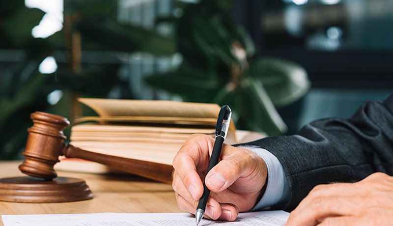 A close-up of a person's hand writing on a document with a pen, with a gavel and stacked law books visible in the blurred background.