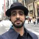 mohammed tayaram - A headshot of a man with a beard and dark fedora hat, wearing a dark jacket and blue headphones, standing in the middle of a pedestrian crosswalk in a busy city street (possibly Ginza, Tokyo), with tall buildings and other people in the background.