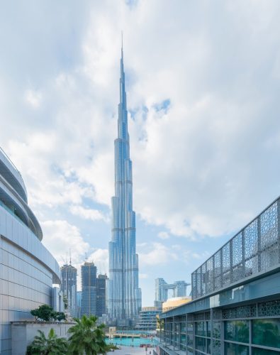 Company formation in the UAE - A wide-angle photo of the Burj Khalifa skyscraper dominating the center of the frame under a partially cloudy blue sky in Dubai. It is viewed from a lower vantage point, flanked by modern buildings with prominent architectural details and metal screens on the left and right, with palm trees and a pool/water feature visible at the base.