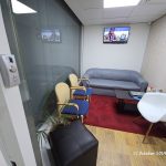stepping stones reception - There is a grey, three-seater sofa centered against the back wall. In front of it is a dark brown/black rectangular coffee table. To the right, there is a white, molded plastic chair with wooden legs, and to the left, two light yellow/beige chairs with blue seats. The seating rests on a deep red area rug.