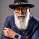 Yusuf Bhai - A stylish portrait of an older man with a long, white beard, wearing a black fedora with a feather band, prominent black-rimmed glasses with orange lenses, a navy blue pinstripe suit, and a wristwatch, posed confidently against a grey background.