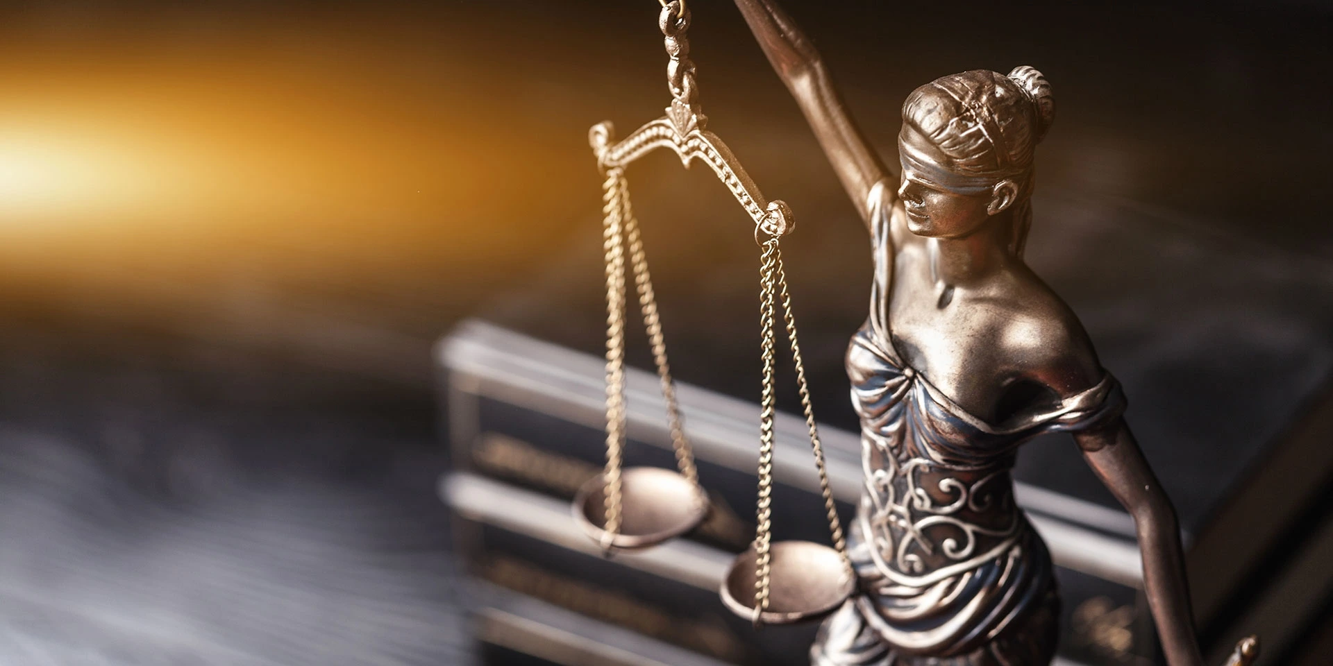 A close-up of a bronze statue of Lady Justice, blindfolded and holding the scales of justice, set against a dark, blurred background with a bright warm light illuminating her from the left.