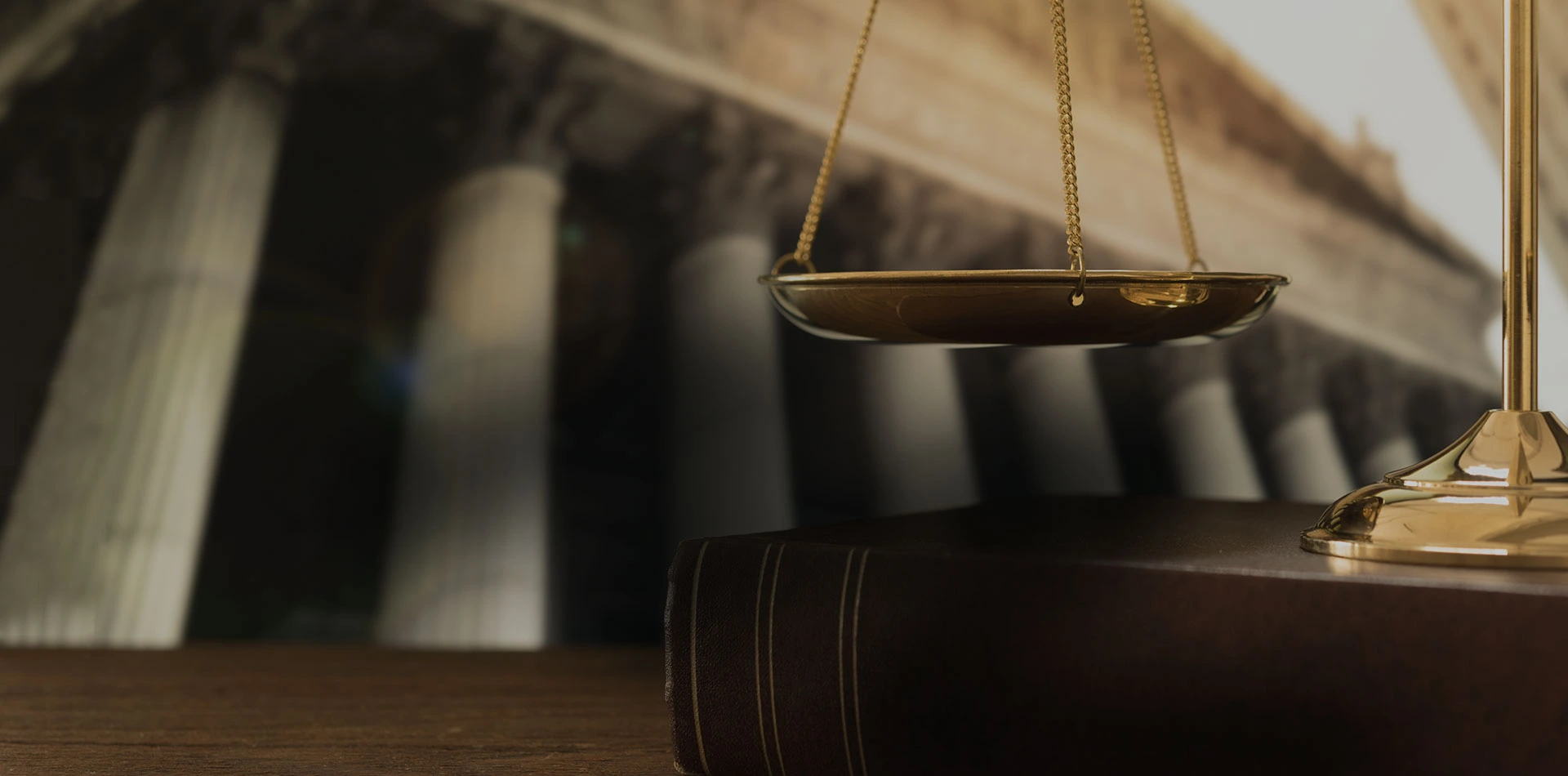 A close-up shot of the brass scales of justice, resting on a thick, dark leather book, with the blurred neoclassical columns of a building in the background.