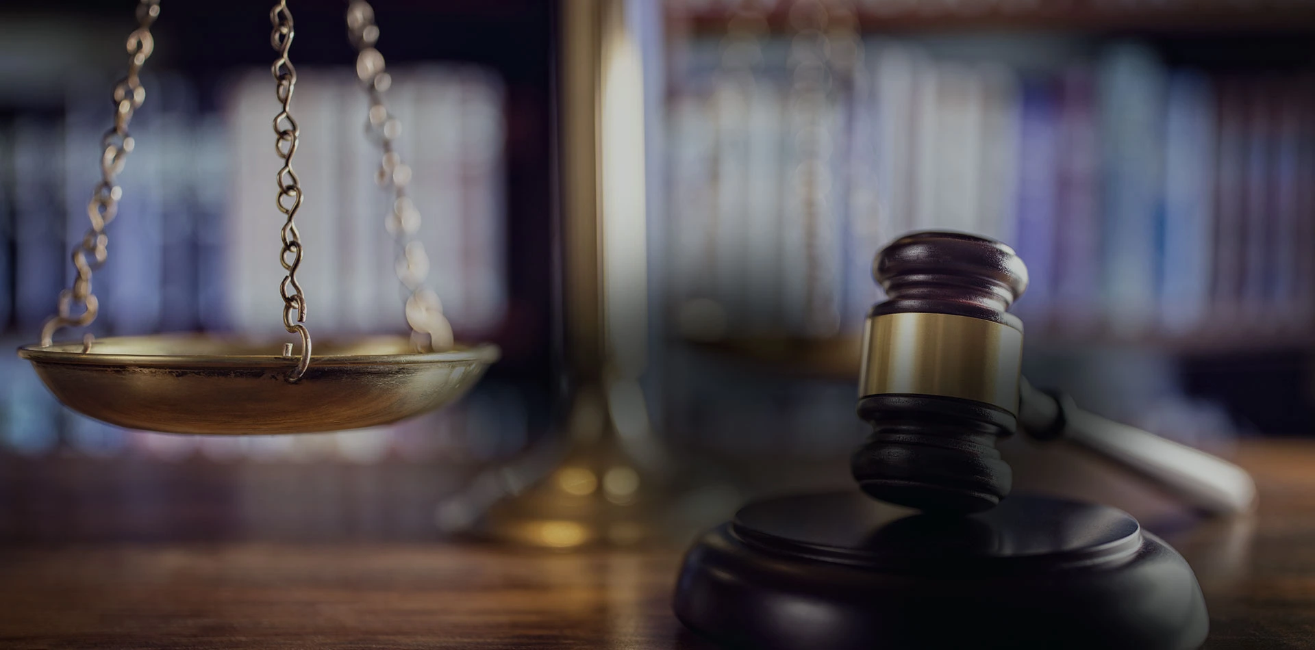 A close-up of a dark wooden gavel with a brass band resting on its sound block on a wooden desk, positioned near the brass scales of justice, with blurred bookshelves in the background.