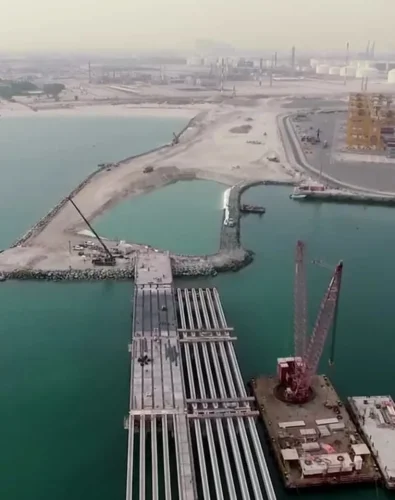 Offshore Company Registration in UAE -Aerial view of a coastal industrial port with pipelines extending over turquoise water toward a dock under construction, cranes and storage tanks visible in the hazy background.